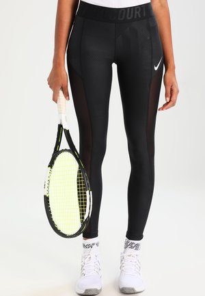 Leggings de sport noirs avec des panneaux en mesh, tissu texturé et taille haute, accompagnés d'une raquette de tennis et de chaussures blanches.