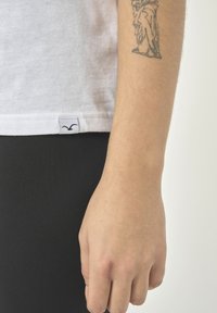 Weißes Baumwoll-T-Shirt mit kleinem Logoschild am Saum, kombiniert mit schwarzen Leggings. Die gezeigte Hand hat ein sichtbares Tattoo.