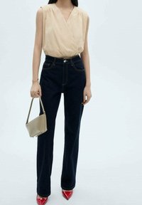 Kvinde iført ærmeløs beige indpakningstop, højtaljede mørke jeans, røde spidse hæle og holder en lille beige håndtaske.