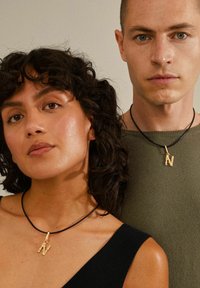 Två individer visar upp guldtonade "N"-halsband på svarta snören, mot en neutral bakgrund. Kvinnans topp är svart, mannens är grön.