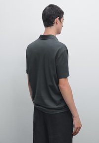 Ung man som står med ryggen vänd, iklädd en mörkgrå t-shirt med kort ärm och svarta byxor, mot en enkel ljus bakgrund.