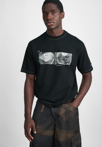 Schwarzes T-Shirt mit kurzen Ärmeln und grafischem Druck auf der Vorderseite. Verfügt über kontrastierende Nähte und einen lockeren Schnitt. Getragen mit braunen gemusterten Cargo-Shorts.
