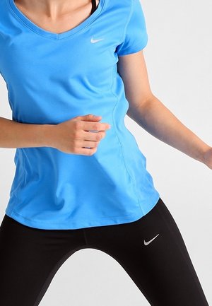 Osoba nosząca niebieską sportową koszulkę Nike i czarne legginsy Nike, w częściowej aktywnej lub rozciągającej pozie na jednolitym białym tle.