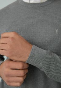 Mâini ajustând manșeta unui pulover tricotat gri purtat peste o bluză albă cu guler, concentrându-se pe textura materialului și poziția mâinilor.