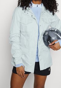 Person i ljusblå jacka och svarta Nike-shorts som håller en svart och grå Nike fotboll i midjehöjd.