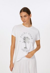 Weißes Baumwoll-T-Shirt mit einem metallischen Palmbaum-Graphic und dem Text "PALMA CLUB". Kurze Ärmel und lockere Passform. Kombiniert mit einem hellen Rock.