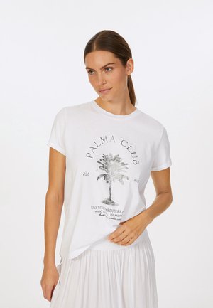 White cotton t-shirt featuring a metallic palm tree graphic and text "PALMA CLUB." Short sleeves and relaxed fit. Worn with a light skirt.