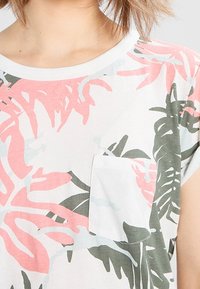 Kortärmad t-shirt i vitt med rosa och grönt tropiskt blommönster, med en liten bröstficka och rund halsringning.