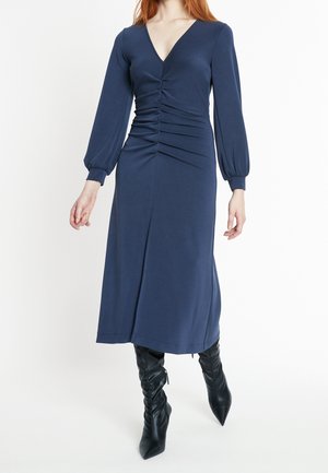 Kobieta ubrana w granatową sukienkę midi z długim rękawem i marszczonym środkowym panelem oraz czarne, luźne buty na szpic, stojąca na białym tle.