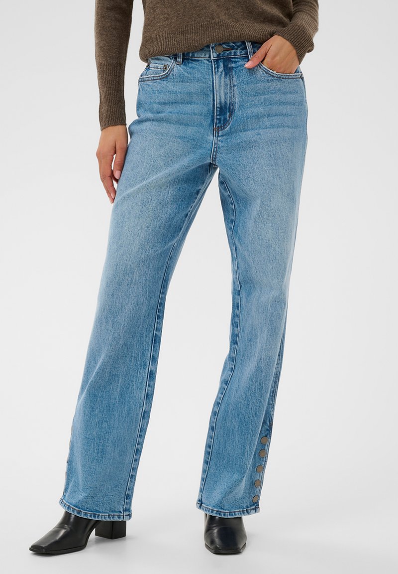 Light blue wide-leg jeans with side snap buttons worn with black heeled boots and a brown long-sleeve sweater, hands in pockets.