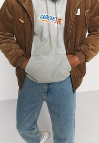 Bruine corduroy zip-up jas met een grijze hoodie eronder met een kleurrafische afbeelding. Lichtblauwe denim jeans en witte sneakers.