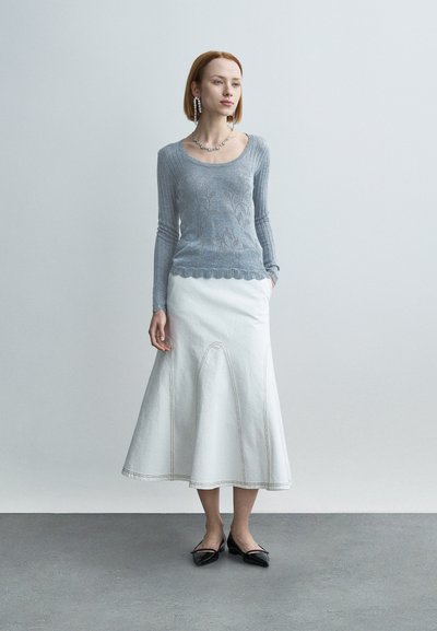 Pull tricoté bleu clair avec motif floral, jupe évasée blanche avec coutures angulaires, et ballerines noires à bout pointu. Accents de bijoux en argent.