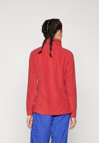 Giacca in pile rossa con colletto alto, cuciture visibili e maniche lunghe, abbinata a pantaloni blu, mostrata da dietro.