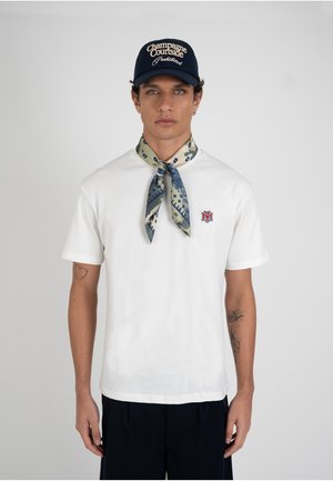 Homme portant une chemise blanche à manches courtes avec un logo rouge, un foulard à motifs autour du cou, un pantalon bleu marine et une casquette bleu marine avec un texte blanc sur un fond uni.