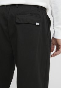 Pantaloni negri din material neted, cu un buzunar din spate cu etichetă și un design pe corp, arătând o parte a unei mâini aproape de talie.