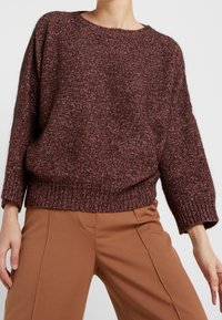 Pull en maille bordeaux à texture épaisse, col rond, épaules tombantes, et ourlet côtelé, associé à un pantalon marron sur mesure.