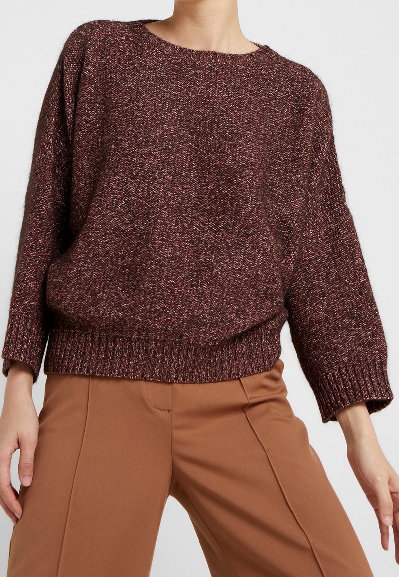 Pull en maille bordeaux à texture épaisse, col rond, épaules tombantes, et ourlet côtelé, associé à un pantalon marron sur mesure.