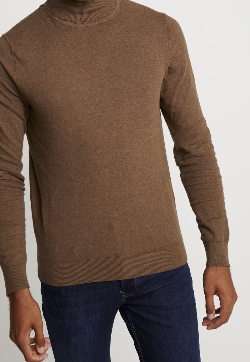 Pull à col roulé brun côtelé en tissu doux et texturé avec des manches longues, assorti à un jean bleu foncé.