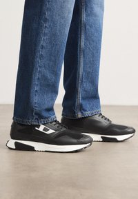 Baskets de sport noires avec un dessus en cuir texturé et en mesh, dotées d'une semelle intermédiaire blanche et d'accents latéraux. Portées avec un jean en denim bleu.