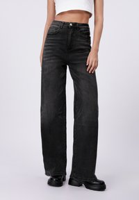 Bredde ben svart denimjeans med høy midje, fem lommer og knappeåpning, kombinert med svarte platåsko. Glatt tekstur.