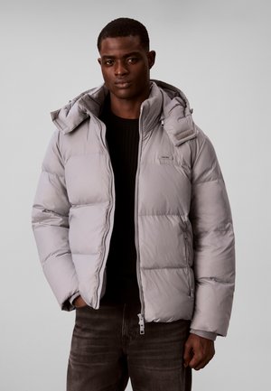 Man wearing a light gray puffer jacket with a high collar and hood, paired with dark jeans and a black sweater.