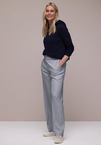 Femme blonde souriante, portant un sweat à capuche bleu marine, un pantalon large à fines rayures gris clair, des baskets blanches, debout les mains dans les poches.