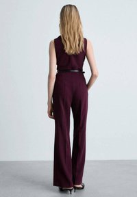 Une combinaison bordeaux avec un corsage ajusté et un pantalon à jambes larges. Stylisée avec une ceinture noire à la taille. Texture lisse, design sans manches.
