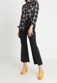Person wearing black and white plaid shirt, black cropped pants with raw hems, and yellow open-toe block heel sandals, standing against white background.