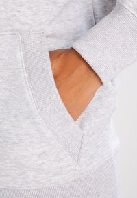 Jasnoszara bluza z kapturem, kangurzą kieszenią i ściągaczami przy mankietach. Miękka faktura z delikatnym melanżowym wzorem. Zbliżenie na dłoń w kieszeni.