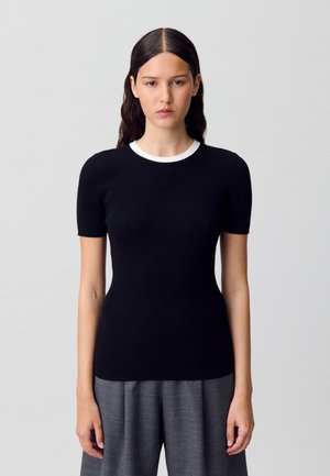 Top corto de punto acanalado negro con cuello redondo blanco y un sutil detalle de texto. Combinado con pantalones de talle alto grises.