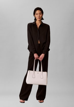 Žena v volné černé košili a kalhotách držící velkou světle béžovou kabelku s dvojitými úchyty na jednobarevném šedém pozadí.