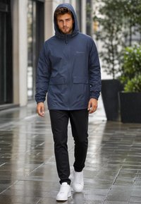 Un homme marche sur un trottoir mouillé, portant une veste à capuche bleu marine, un pantalon noir et des baskets blanches dans un cadre urbain extérieur.