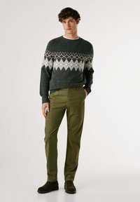 Šedý vzorovaný sveter s krátkymi rukávmi, so diamantom v bielej a oranžovej farbe, spárovaný s priliehavými olivovozelenými nohavicami a hnedými čižmami.