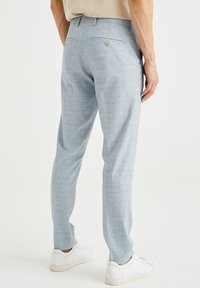 Muž oblečený v světle modrých kostkovaných kalhotách a bílých teniskách, stojící s uvolněnýma rukama po stranách na bílém pozadí.