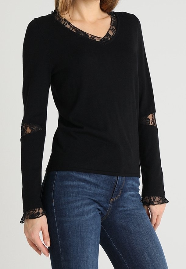 Femme portant un haut noir à manches longues avec des détails en dentelle sur le décolleté, les manches et les poignets, associé à un jean bleu.