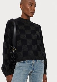 Pull à carreaux noir et doré avec un col haut, coupe ample, texture tricotée et ourlet côtelé, associé à un jean bleu et un sac à main.