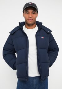 Navy puffer jacket with a zip front, large collar, and logo patch. Worn over a white shirt, with blue jeans and a dark cap. Textured fabric.