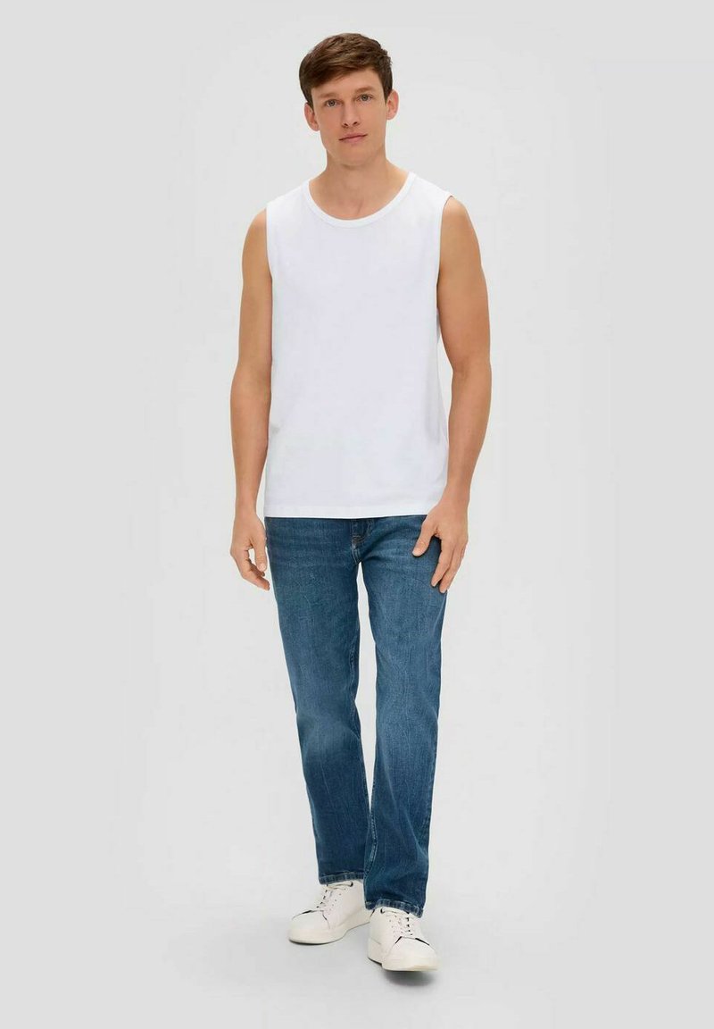 Young man standing wearing white sleeveless shirt, blue jeans, and white sneakers against a plain light background.