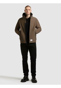 Brązowa kurtka zapinana na zamek z wysokim kołnierzem i polarem w środku, w zestawieniu z czarnymi spodniami i czarnymi butami. Materiał wydaje się gładki, o dopasowanym kroju.