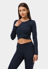 Merine sinise pika varrukaga lühendatud topp, millel on keerutamisdisain, koos kõrge vöökohaga sobivate retuusidega. Veniv, sile kangas koos peene logo detailiga.