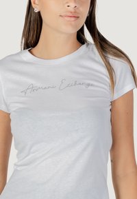 Witte katoenen T-shirt met korte mouwen, voorzien van zilverkleurige geborduurde tekst "Armani Exchange" over de borst. Gladde textuur.