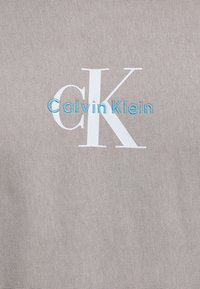 Tessuto grigio con testo bianco "CK" e blu "CalvinKlein", caratterizzato da una texture liscia e un design minimalista.