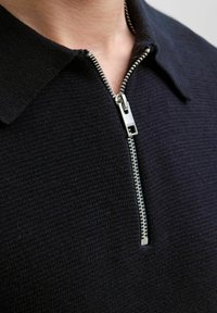 Tuvas tuvplāksne ar personu, kas valkā tumšas adītas polo krekls ar daļēji atvērtu sudraba rāvējslēdzēju apkaklē.