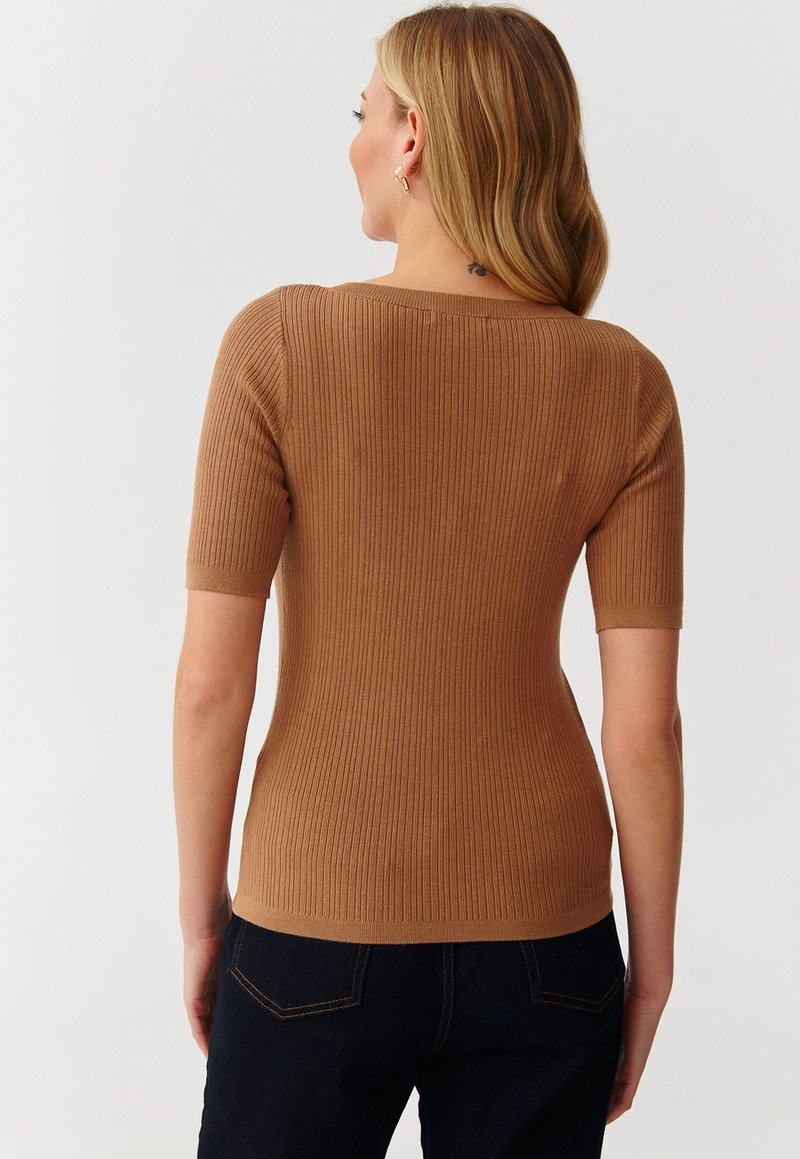 Prążkowana bluzka krótki rękaw w jasnobrązowym kolorze; dopasowany krój z okrągłym dekoltem i teksturowanym materiałem. Połączona z ciemnymi dżinsami.