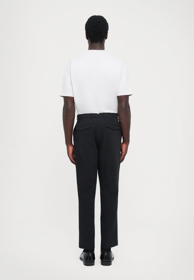 Tricou alb din bumbac și pantaloni negri cu buzunare. Pantalonii au un croi drept și sunt purtați cu pantofi negri. Vedere din spate.