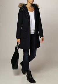 Parka bleu marine avec capuche à bord en fausse fourrure, fermeture éclair frontale et deux grandes poches. Des bottines noires en cuir à la cheville et un sac cabas noir sont inclus.