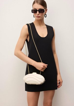 Mujer con vestido negro sin mangas y gafas de sol rectangulares negras, sosteniendo un bolso crema con forma de concha y cadena dorada.