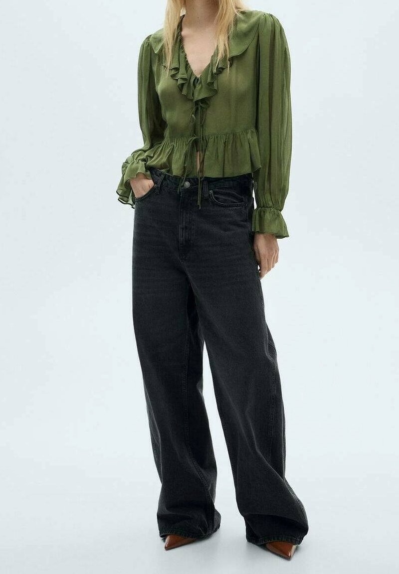 Frau in einer transparenten, olivgrünen, rüschierten Bluse mit langen Ärmeln und lockeren, schwarzen weit geschnittenen Jeans, die mit einer Hand in der Tasche steht.