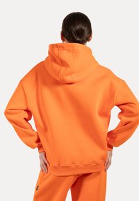 Sudadera oversized naranja con capucha ajustable, puños y dobladillo acanalados, fabricada en una tela suave. Mostrada desde la parte de atrás con un corte relajado.