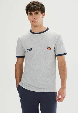 Hombre con camiseta gris claro con ribete y logos azul marino, combinado con pantalones azul marino, de pie frente a un fondo claro y liso.